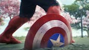 Captain America's shield is embedded in the pavement after a big impact. standing behind it, we see the legs of the Red Hulk